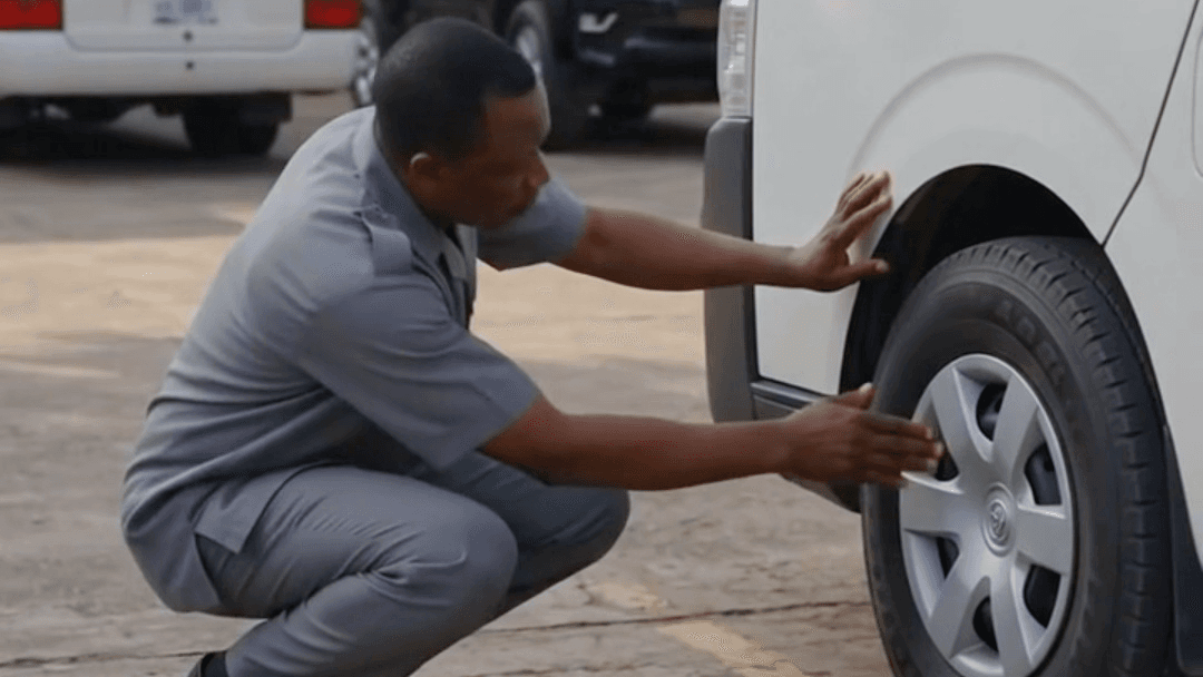 A driver crouching to check the tyre of a Toyota Hiace in a government office compound.